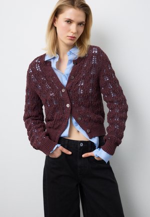 Giovane donna che indossa una camicia blu con bottoni sotto un cardigan lavorato a maglia color vinaccia, abbinata a pantaloni neri a vita alta, con le mani nelle tasche.