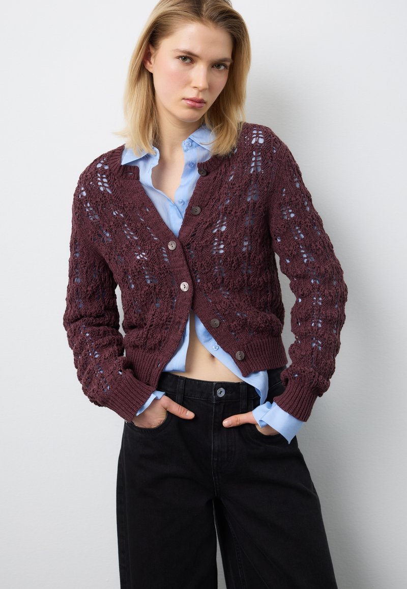 Junge Frau trägt ein blaues Hemd mit Knöpfen unter einer bordeauxroten Strickjacke, kombiniert mit hoch taillierten schwarzen Hosen, Hände in den Taschen.