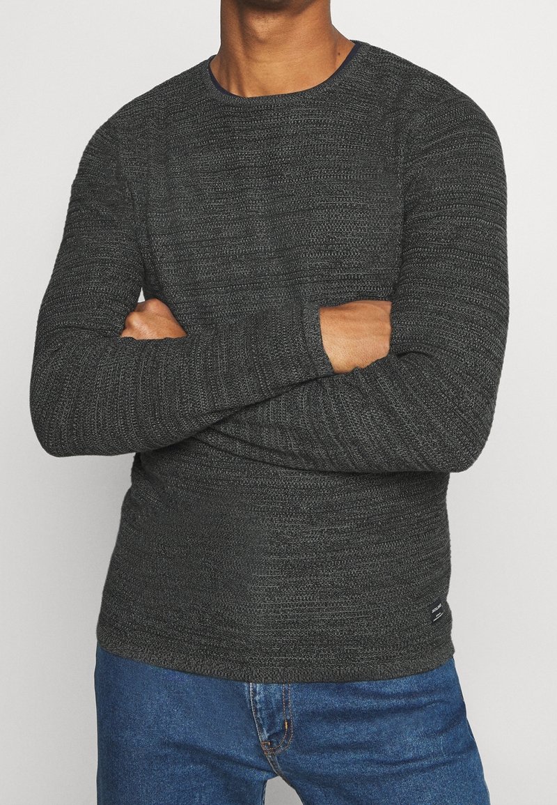 Homme portant un pull texturé gris foncé, les bras croisés, associé à un jean en denim bleu, debout devant un fond clair uni.