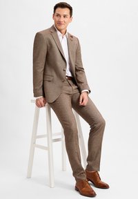 Brauner strukturierter Anzug mit tailliertem Schnitt, weißem Hemd und braunen dressierten Schuhen. Schmale Hosen und zwei Knöpfe am Sakko.