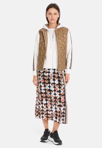 Quilted brown vest over a white hoodie, paired with a long black skirt featuring a hearts pattern. Black athletic shoes complete the outfit.