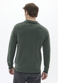 Polo shirt cu mâneci lungi, de culoare verde închis, cu guler, manșete rigate și tiv drept. Textură moale, detalii minime, purtat cu blugi gri.