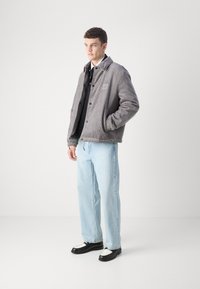 Grå kordfløyelsjakke med krage og logodetalj, båret over en svart skjorte, parret med lyseblå jeans og svarte sko med hvite detaljer.