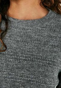 Gros plan sur une personne portant un pull gris texturé en maille avec un col rond et des cheveux bouclés brun clair épais.