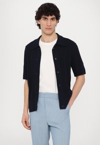 Sini lyhythihainen nappipaita, jossa on muotoiltu malli, yhdistettynä vaaleansinisiin raidallisiin housuihin. Valkoinen pyöreäkaulainen t-paita alla.