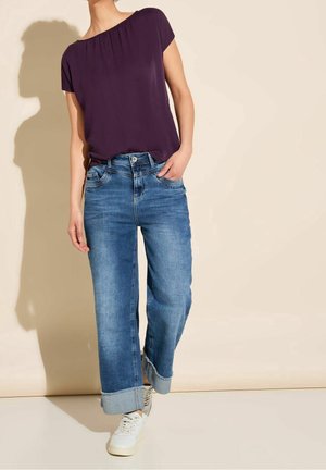Persoon die een paarse blouse met korte mouwen, opgerolde blauwe spijkerbroek en witte sneakers draagt, staand tegen een lichtbeige achtergrond.