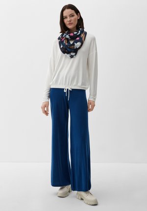 Frau trägt ein weißes langärmeliges Oberteil, blaue Hose mit weitem Bein, beige Plateauschuhe und einen mehrfarbigen Blumen-Schal, stehend vor weißem Hintergrund.