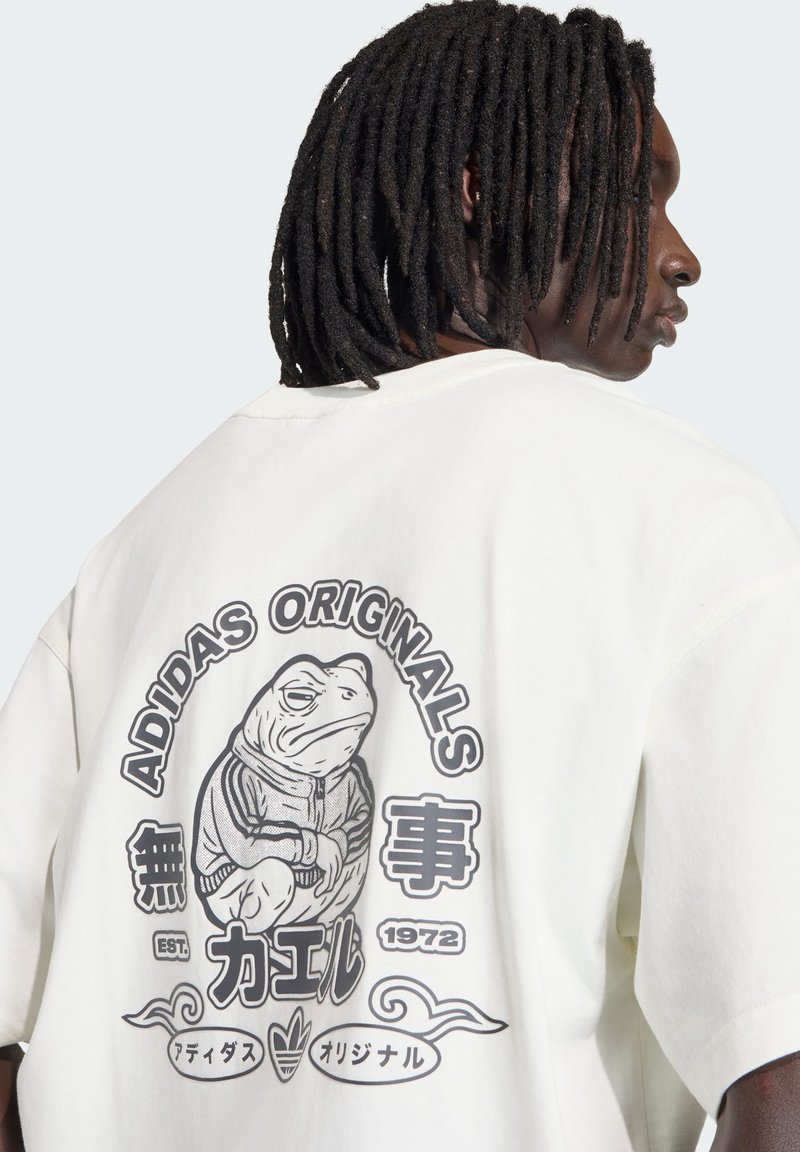 T-shirt oversize couleur crème avec un motif de grenouille gris, agrémenté de texte. Coupe décontractée, texture douce et manches courtes.