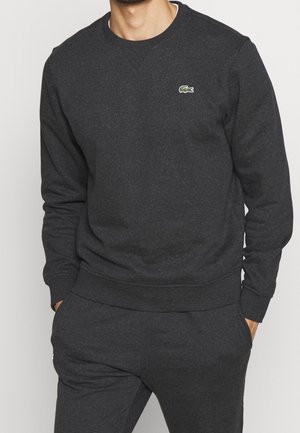 Mand iført mørkegrå crewneck sweatshirt og matchende sweatpants med hænderne i lommerne, viser lille grønt krokodillelogo på brystet.