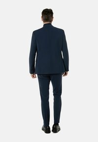 Marine blazer en bijpassende broek, slim fit, gladdige stof, enkele split aan de achterkant, gedragen met zwarte schoenen. Zichtbare naaddetails.