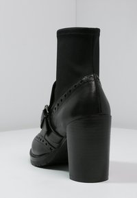 Bota de cuero negra hasta el tobillo con un upper de tela elástica, tacón grueso, acento perforado y un detalle de hebilla lateral. Textura suave en general.