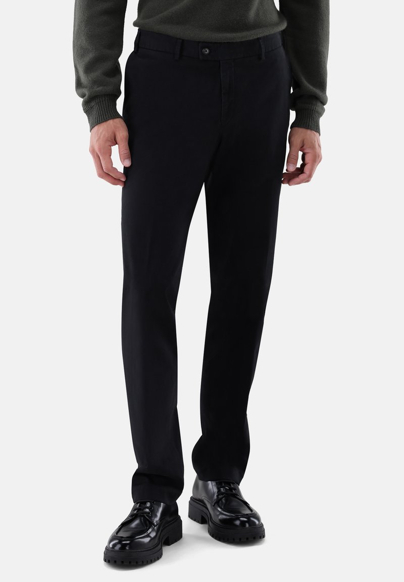 Schwarze Hosen aus glattem Stoff mit schmal zulaufender Passform, ausgestattet mit einem Knopfverschluss und seitlichen Taschen. Kombiniert mit schwarzen, klobigen Schuhen.