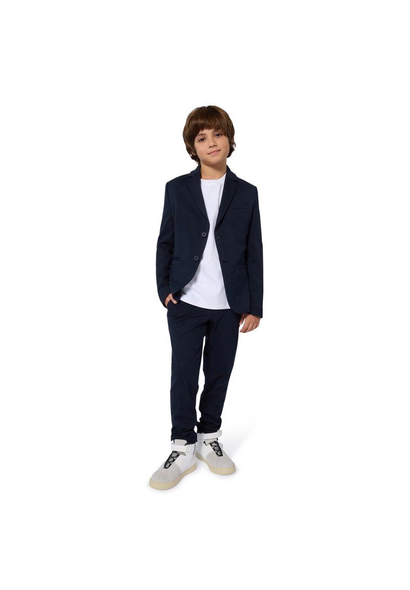 Tengerészkék öltöny, testhezálló blézerrel és hozzáillő nadrággal, fehér pólóval és világos színű tornacipővel párosítva. Simább anyag, modern szabás.