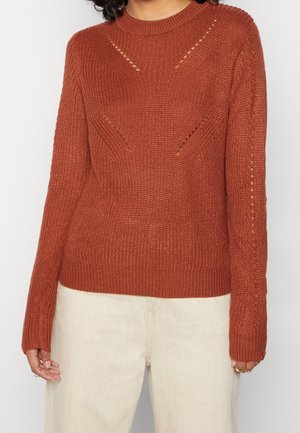 Rust-colored knitted sweater with subtle open stitch patterns, worn with light beige pants on a person with curly hair.