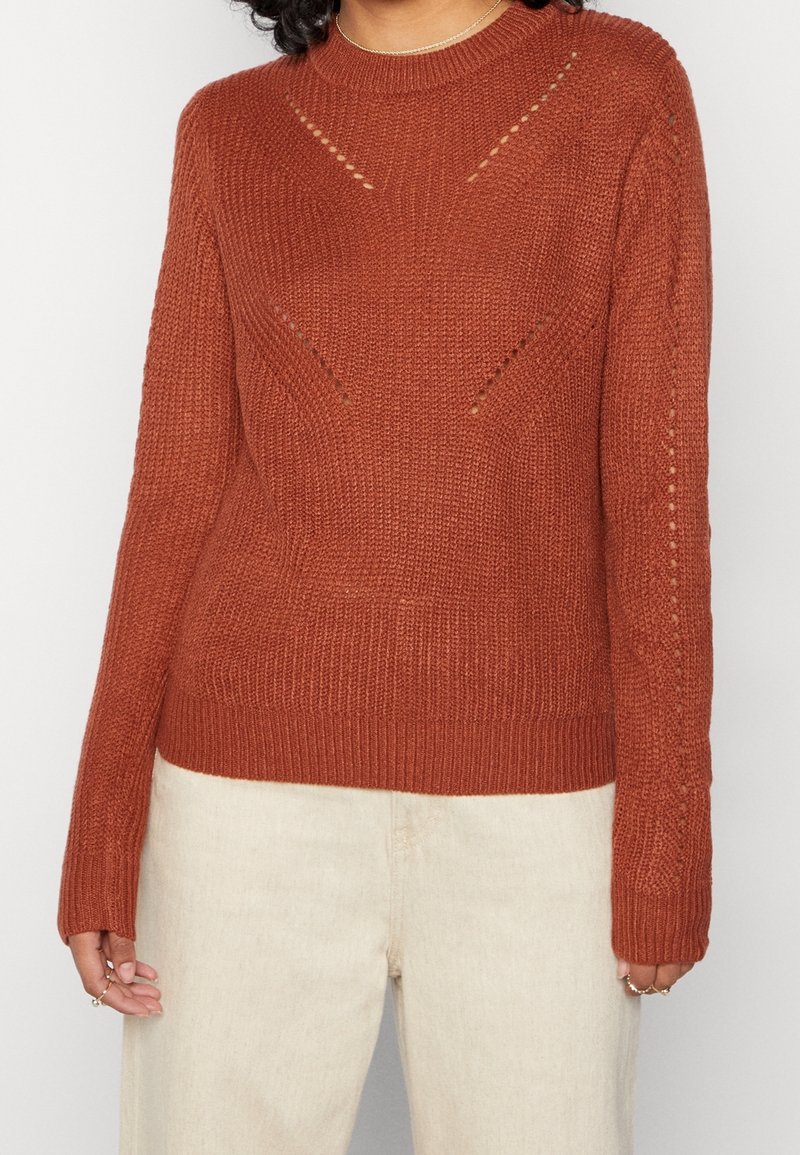 Pull tricoté couleur rouille avec des motifs délicats en points ajourés, porté avec un pantalon beige clair sur une personne aux cheveux bouclés.