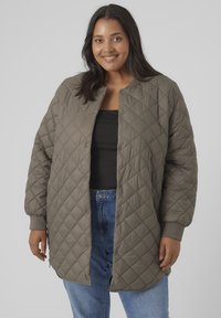 Quilted dark olive green jacket, round neck, snap buttons, ribbed cuffs, long sleeves, and side slits, worn over a black top and denim jeans.