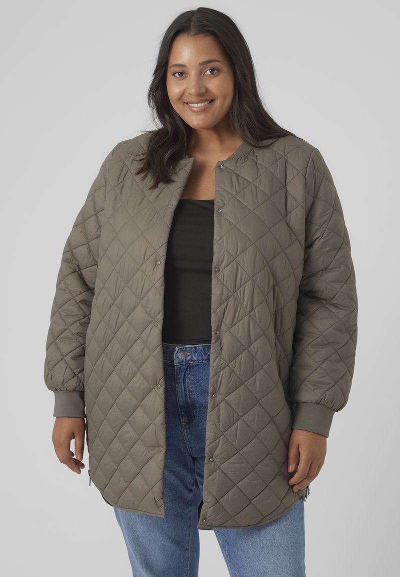 Quilted dark olive green jacket, round neck, snap buttons, ribbed cuffs, long sleeves, and side slits, worn over a black top and denim jeans.