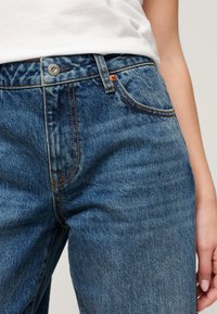 Dżinsy w średnim niebieskim odcieniu z pomarańczowymi szwami, pięć kieszeni, metalowy guzik w pasie. Gładka tekstura, prosty krój.