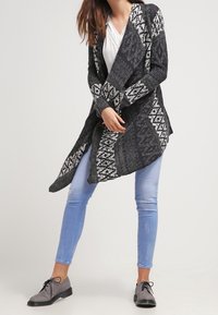 Cardigan de punto en gris oscuro con patrones geométricos blancos, de abertura frontal, mangas largas y un diseño suelto y fluido. Lleva puesto un top blanco.