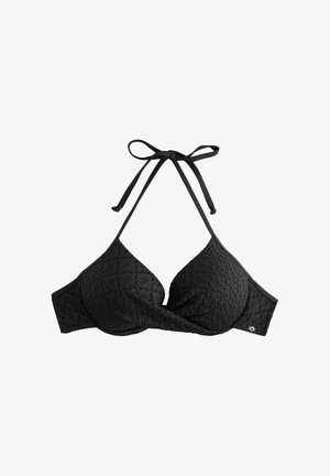 Donna in bikini nero in piedi su una spiaggia sabbiosa con acqua blu e colline verdi sullo sfondo sotto un cielo sereno.