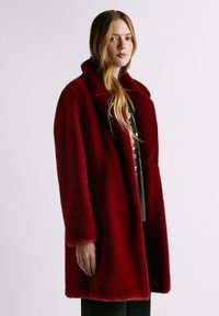 Cappotto in pelliccia sintetica rossa con colletto largo, maniche lunghe e vestibilità comoda. La texture appare morbida e vellutata, aumentando la sensazione di calore complessiva.