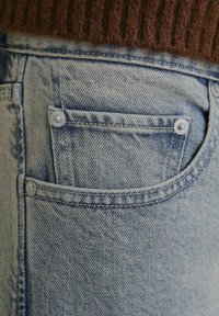 Tela de mezclilla de color azul claro que muestra un primer plano de un bolsillo con herrajes plateados y detalles de costura. La textura está ligeramente desgastada.