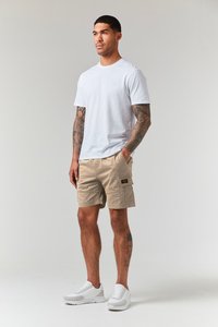 Weißes Baumwoll-T-Shirt, beigefarbene Cargo-Shorts mit Taschen und weiße Sneaker. Hat Tattoos an den Armen; neutraler Hintergrund.