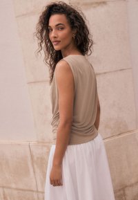 Haut beige sans manches avec un détail froncé à la taille, accompagné d'une longue jupe blanche. En arrière-plan, un mur clair texturé.