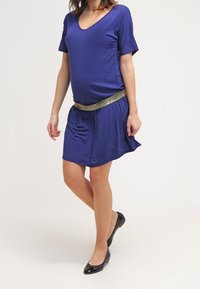 Lyhythihainen V-mallinen mekko syvänsinisestä kankaasta, jossa on ryhditetty vyötärö ja kultainen metallinen vyökoriste. Yhdistetty mustiin ballerinoihin.