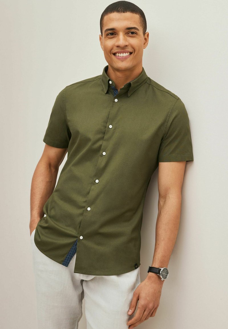 Camisa de manga corta en color verde oliva con botón frontal, textura sutil y tela de patrón azul en contraste en la solapa interior.