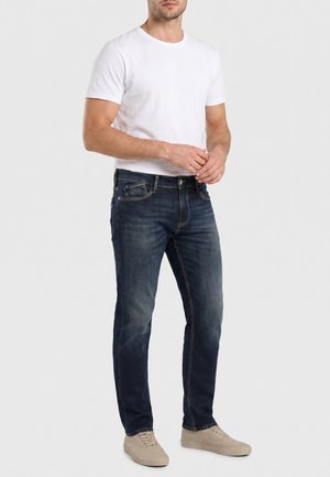 Hombre con camiseta blanca de manga corta, vaqueros azul oscuro con costuras visibles y zapatillas beige claras, de pie frente a un fondo liso.