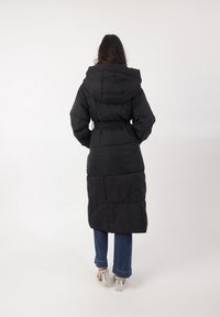 Mujer con cabello largo y oscuro de pie de espaldas, vistiendo un abrigo acolchado negro con cinturón, jeans azules y sandalias de tacón bloque transparente, sobre fondo blanco.