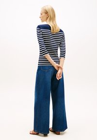 Μπλε ναυτικό και λευκό ριγέ μακρυμάνικο τοπ σε συνδυασμό με φαρδιά μπλε τζιν παντελόνια. Το μοντέλο στέκεται με τα χέρια σταυρωμένα πίσω από την πλάτη. Ομαλές υφές.