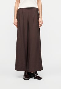 Femme portant un pantalon large marron foncé et des bottines à talons marron foncé, debout devant un fond clair uni.