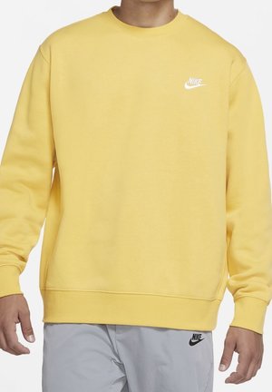Mężczyzna w żółtej bluzie Nike z białym logo i jasno szarych spodniach Nike z czarnym logo, stojący na białym tle.