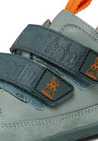 Scarpa in pelle verde con cinturini in tessuto grigio, caratterizzata da un logo di coniglio arancione sul cinturino. Il design presenta texture contrastanti e una forma arrotondata.
