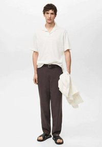 Polo bianco, pantaloni marrone scuro, sandali neri. Il modello tiene una maglietta beige chiara. Vestibilità rilassata, maniche corte, texture minima.