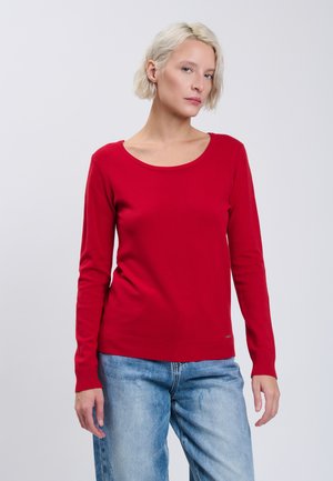 Femme blonde aux cheveux courts portant un pull rouge à manches longues et un jean bleu clair, debout devant un fond gris clair.