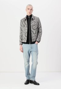 Gråmønstret jakke med lynlås over en sort turtleneck, parret med lyseblå jeans og sorte sko, sat mod en hvid baggrund.