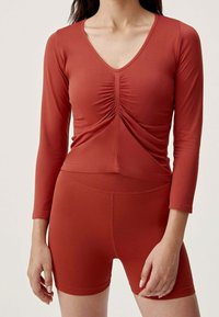 Red long-sleeve top with a V-neck and gathered detail at the front, paired with matching red fitted shorts. Smooth fabric with slight sheen.