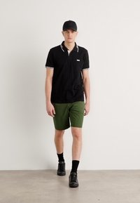 Polo noir avec bord blanc et logo, associé à un short vert. Le modèle porte des baskets noires et une casquette assortie, debout sur un sol neutre.