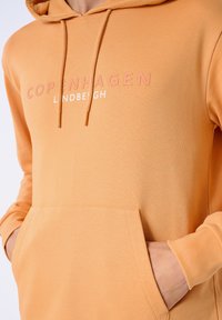 Orange hoodie med känguruficka, med "COPENHAGEN" och "LANDBEIGH" tryckt i vitt och rosa. Mjuk, texturerad tyg.