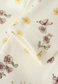 Zbliżenie lekkiej, kwiecistej tkaniny w kolorze kremowym, z małymi fioletowymi i żółtymi kwiatami, z widoczną etykietą marki.