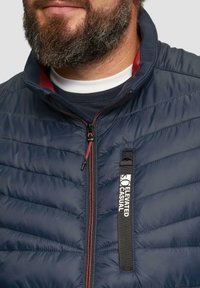 Tamnoplava jakna s puhanjem koja ima visoki ovratnik, crvene detalje, potpuni zatvarač i oznaku 'Elevated Casual'. Glatka tekstura.