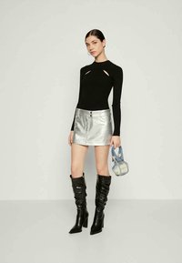 Haut noir à manches longues avec détails découpés, mini jupe argentée avec boutons sur le devant, bottes noires brillantes montant jusqu'aux genoux, et petit sac à main bleu.