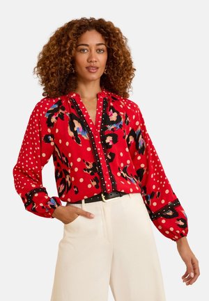Frau mit lockigem Haar, die eine rote Bluse mit Polka-Dots und abstrakten Mustern trägt, kombiniert mit weißen hoch taillierten Hosen und einem schwarzen Gürtel.