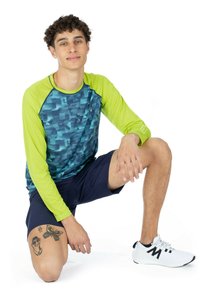 Langarm zwemshirt in blauw en neongroen, met een geometrisch patroon. Draagt een donkere korte broek en witte sportschoenen.