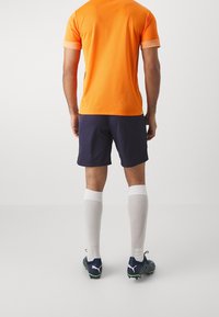 Man med orange sporttröja, marinblå shorts, vita knähöga strumpor och marinblå fotbollsskor står vänd bort från kameran mot en enfärgad bakgrund.