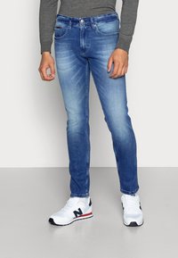 Světlé modré džíny z denimu s úzkým střihem, vybavené popraskaným designem, pěti kapsami a zapínáním na knoflík, nošené s bílými sportovními botami.