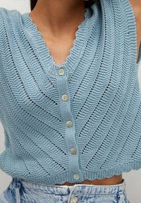 Lichtblauw gebreid crop top met een V-hals, voorzien van een chevronpatroon, vijf knopen aan de voorkant en een getextureerde geribbelde afwerking. Zacht katoenen materiaal.